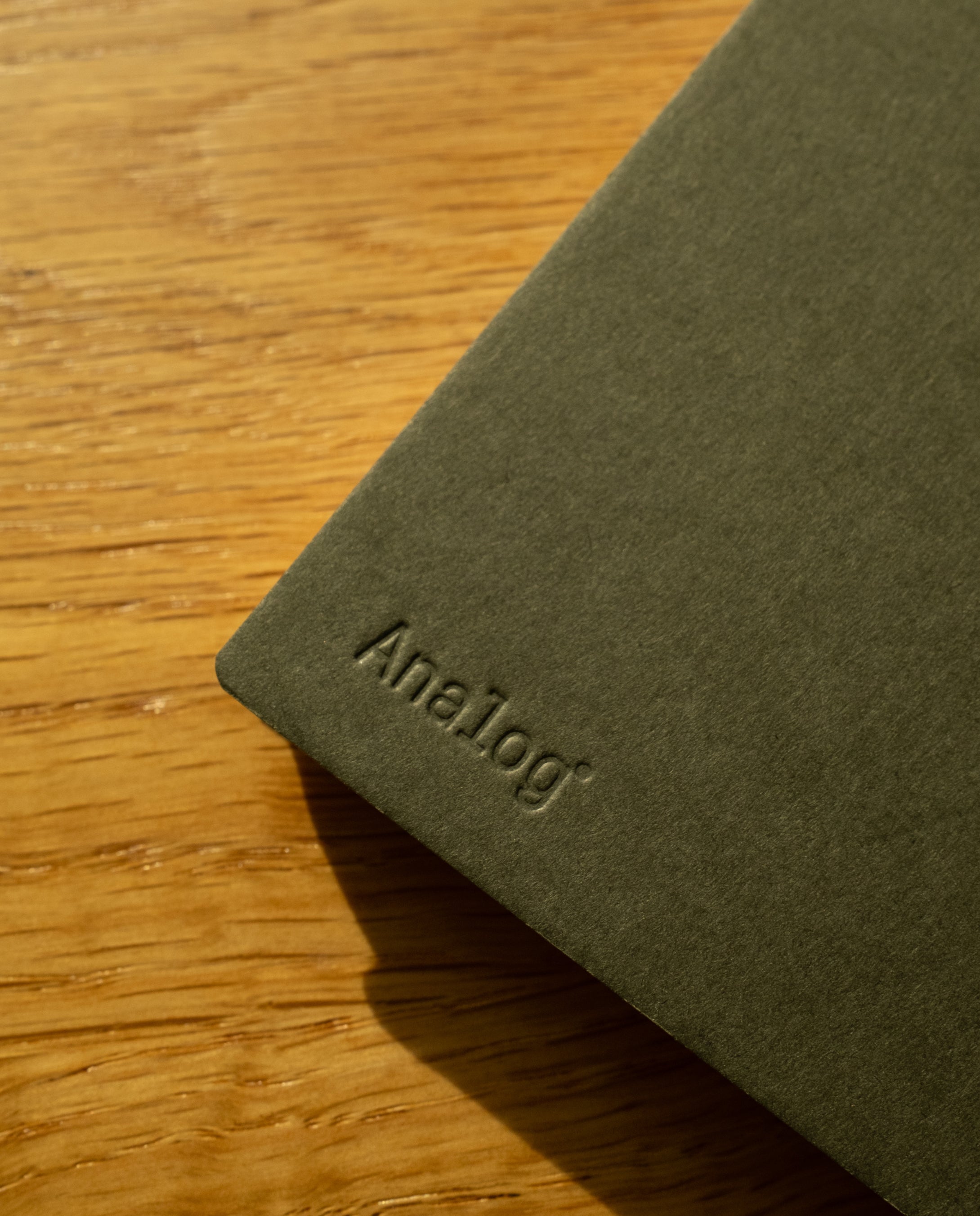 A Discbound Analog + 3 Refills notebook with brass disc binding rests on a wooden surface, soft sunlight highlighting the dark green cover and textured materials.
