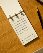 The Discbound Analog + 3 Refills, a compact notepad with brass discs, sits on a wooden desk beside a black and gold pen, open to a handwritten to-do list for creative tasks, fabric swatches, photoshoots, and calls.