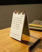 The Discbound Analog + 3 Refills, a compact refillable planner for handwritten tasks, to-do lists, and scheduled calls with brass disc binding, rests on a wooden table beside a pen and book in warm sunlight.