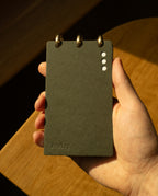A hand holds a small, dark gray Discbound Analog + 3 Refills notepad with brass disc binding at the top. The cover has three white dots and "Analog" embossed, lit warmly with soft shadows on the hand and wooden surface.