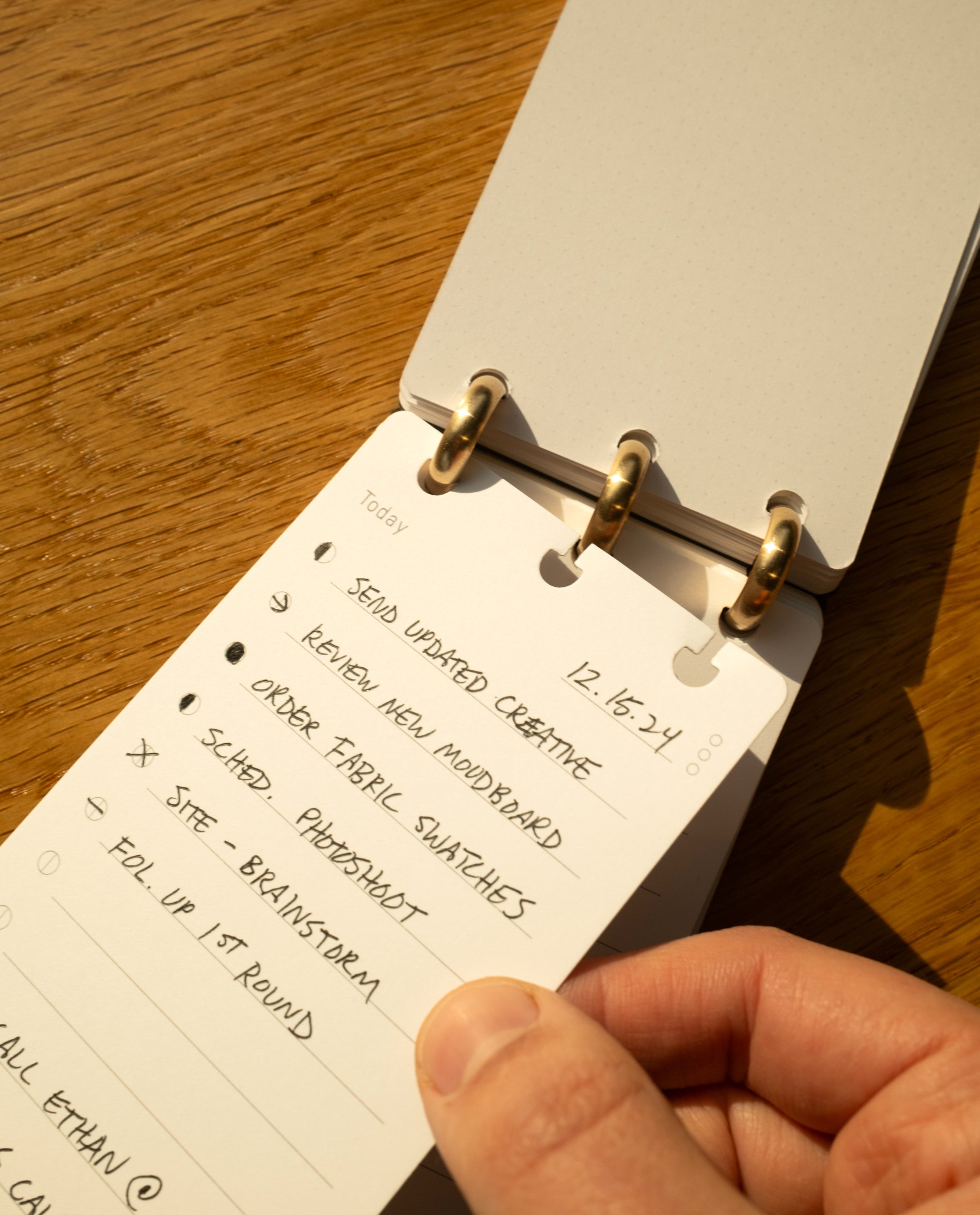 A hand holds a small notepad with a to-do list, featuring tasks like sending updated creative, removing an old moodboard, and ordering fabric swatches. Sunlight casts warm shadows on the wooden desk below, where a Discbound Analog Refill (1-Pack) sits neatly beside the notepad.