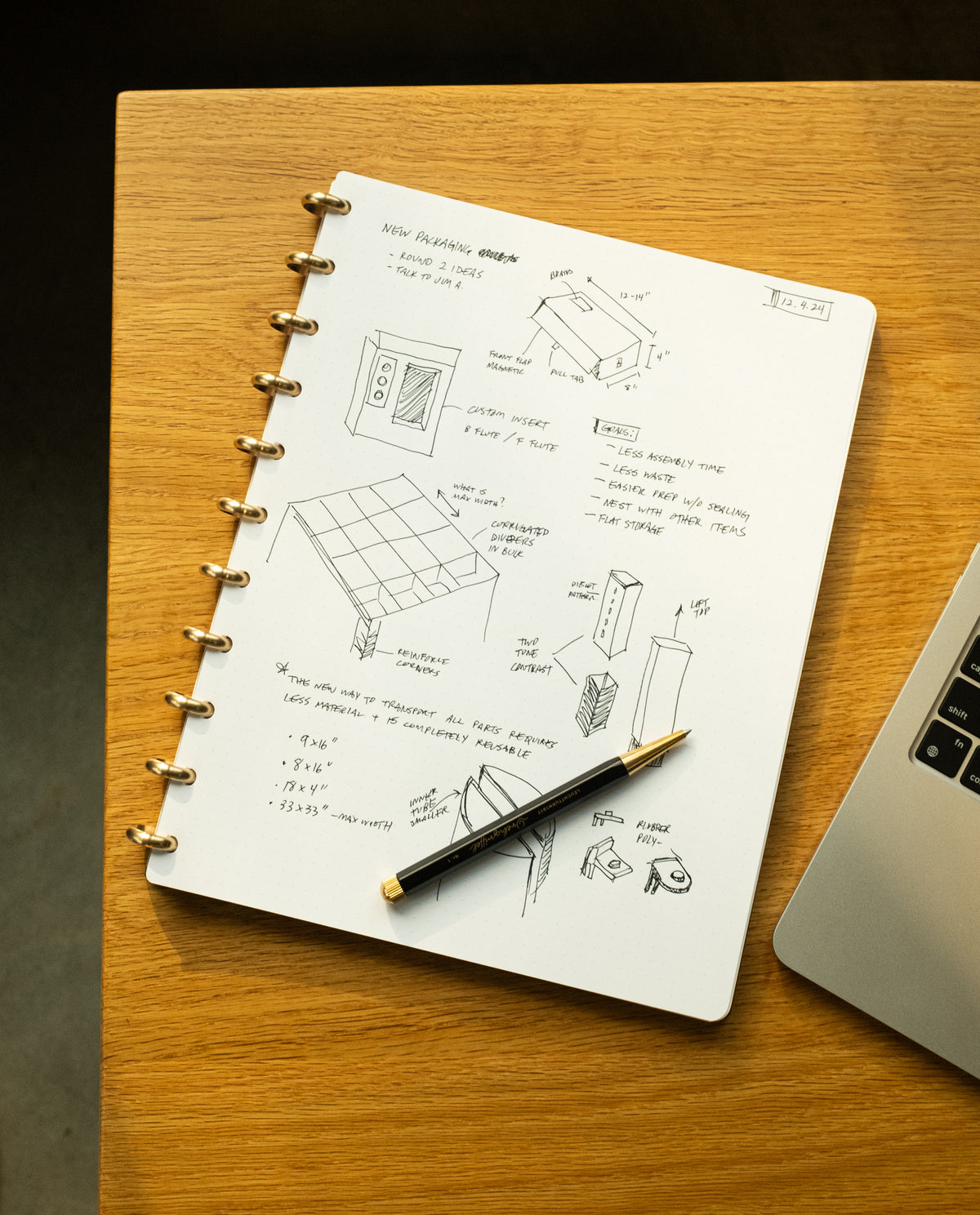 The Discbound Journal Bundle (8.5" x 11" Journal + 3 Refills), filled with sketches, handwritten notes, and diagrams with measurements, rests on a wooden desk beside a fountain pen and the edge of a laptop. Warm lighting casts a glow on the journal’s paper, brass discs, and the smooth surface of the desk.