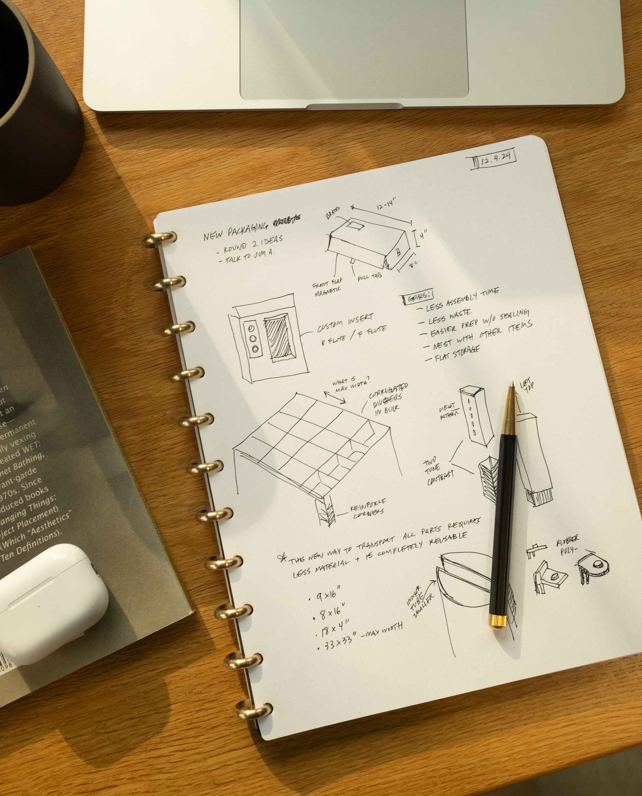 An open Discbound Journal Bundle (8.5" x 11" journal + 3 refills) filled with sketches and notes sits on a wooden desk, accompanied by a black pen resting on top, a coffee cup, a book, and a white wireless earbuds case. Sunlight streams across the scene, highlighting the journal’s elegant brass discs that lend an air of sophistication to this creative workspace.