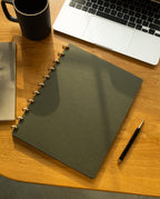 A Discbound Journal Bundle (8.5" x 11" journal with three refills) featuring elegant brass discs, alongside a black pen, open laptop, and black mug, sits on a wooden desk bathed in warm sunlight—creating a cozy and productive workspace atmosphere.
