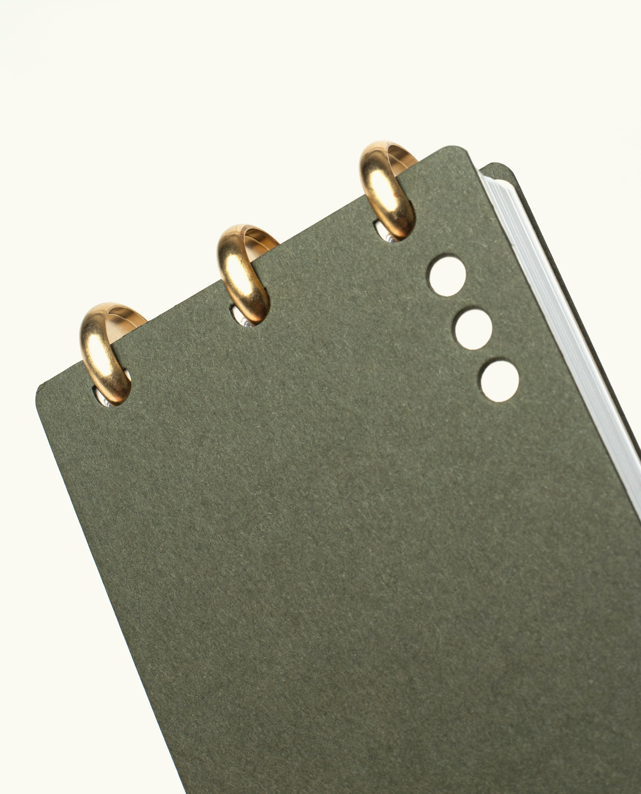 A close-up of the Discbound Analog + 3 Refills, a dark green brass disc-bound notebook with three small round holes on the cover, shown on a plain white background—a refined, refillable task planner.