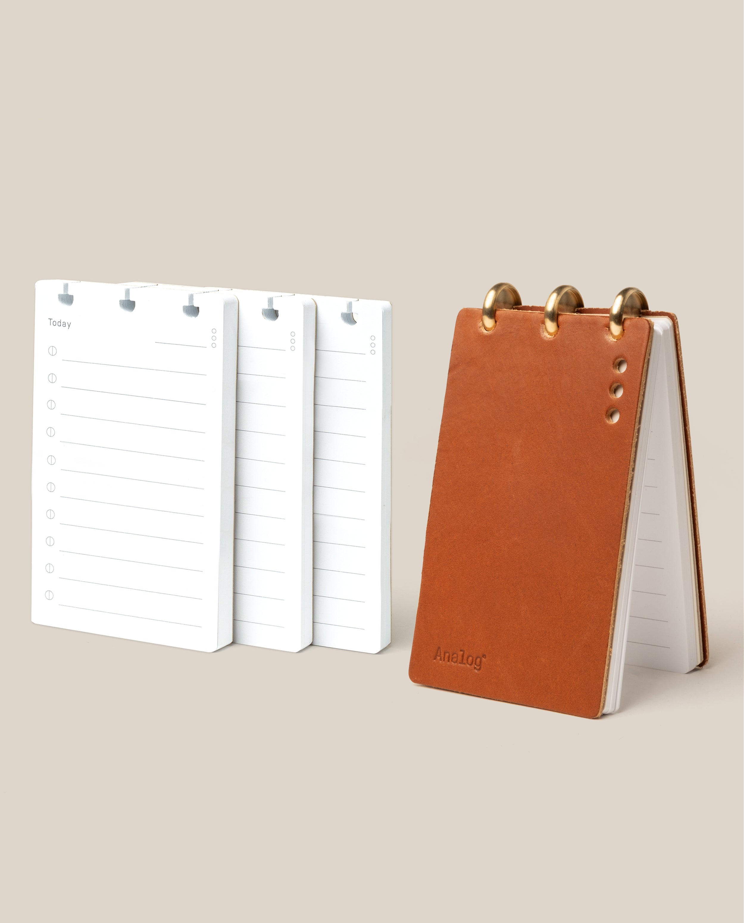 The Leather Discbound Analog + 3 Refills (Tan) features a tan full-grain leather notepad with gold disc rings, shown standing upright beside three stacks of loose refills on a beige background.