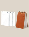 The Leather Discbound Analog + 3 Refills (Tan) features a tan full-grain leather notepad with gold disc rings, shown standing upright beside three stacks of loose refills on a beige background.