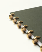 A close-up of the Discbound Journal Bundle (Standard 6"x 8.5" Journal + 3 Refills) featuring a refillable dark green cover and large gold-colored disc binding rings on a white background.