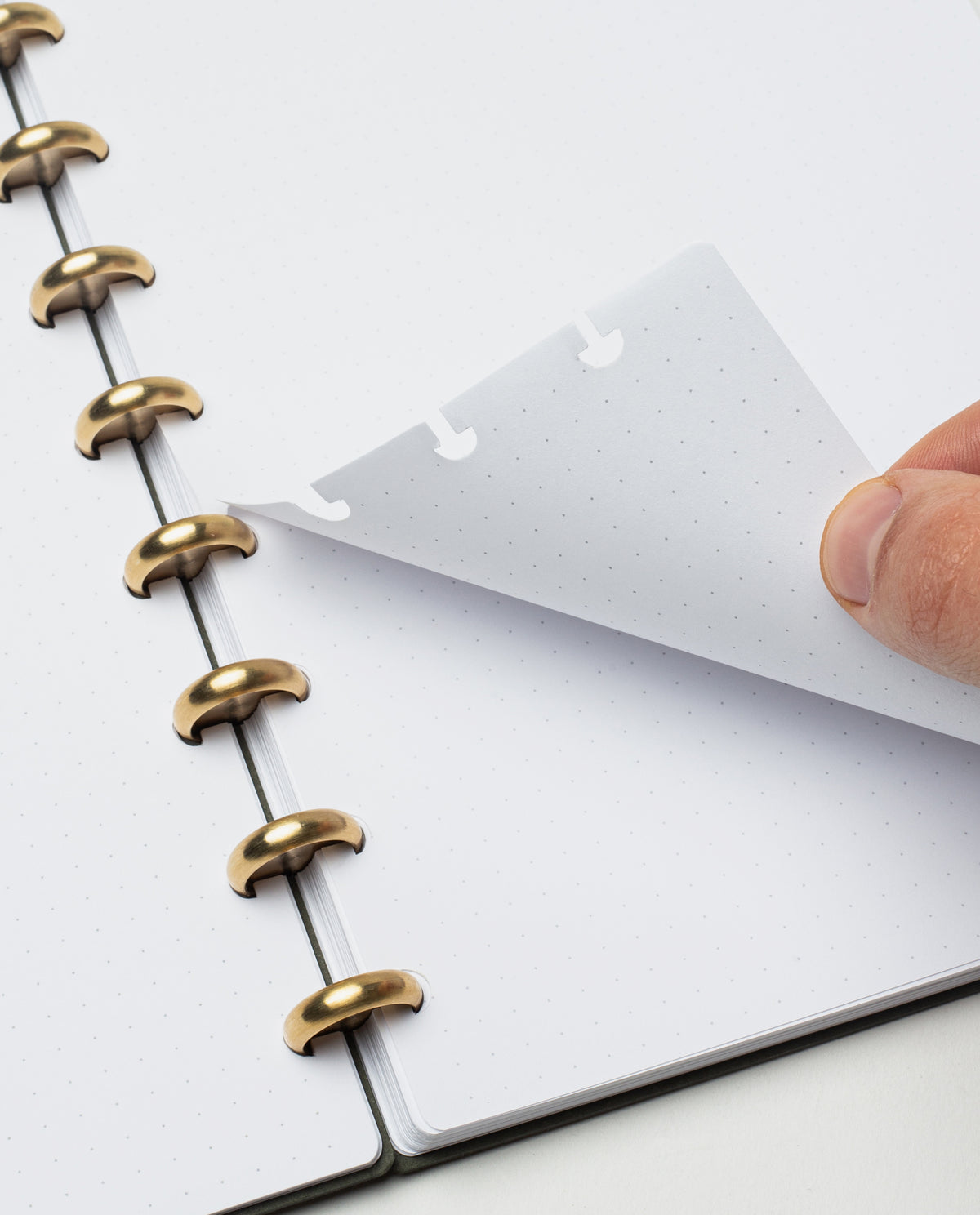 A hand lifts a sheet of dot grid paper from the Discbound Journal Refill (Large 8.5" x 11" - 3 Pack) with elegant gold rings. The background is white, and the notebook pages are blank.