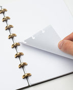 A hand lifts a dotted page in the Discbound Journal (Standard 6"x 8.5") with solid brass discs. The refillable journal rests open on a white surface, showing blank pages with light gray dots.