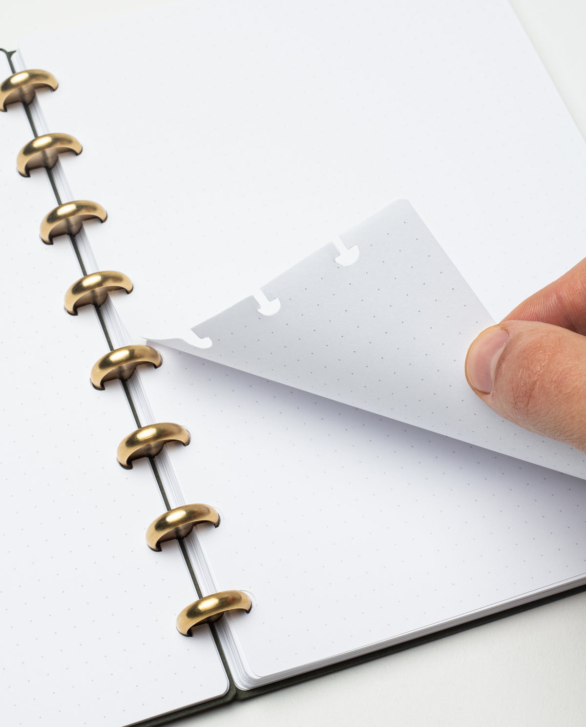 A hand lifts a dotted page in the Discbound Journal (Standard 6"x 8.5") with solid brass discs. The refillable journal rests open on a white surface, showing blank pages with light gray dots.