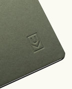 A close-up of the corner of the Discbound Journal (Standard 6"x 8.5") in green, featuring an embossed geometric logo on the cover and solid brass discs binding partially open white pages inside.