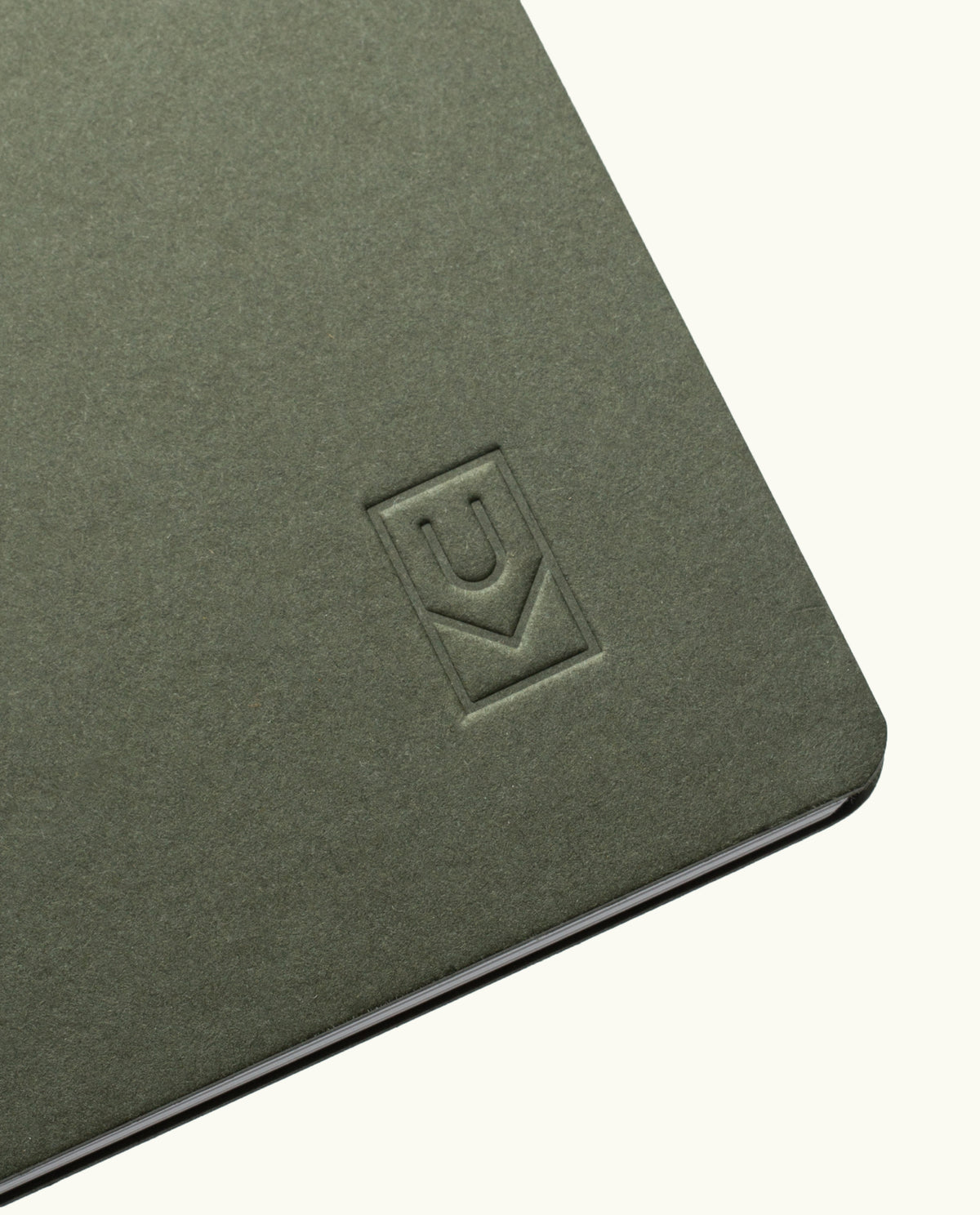 A close-up of the corner of an olive green Discbound Journal Bundle (8.5" x 11" Journal + 3 Refills) with a debossed logo featuring a U and a check mark inside a rectangle.