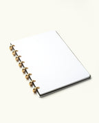 A Discbound Journal (Standard 6"x 8.5"), featuring a white dotted interior, black cover, and solid brass discs, rests on a light plain surface.