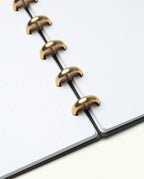 An open Discbound Journal (Standard 6"x 8.5") with white dotted pages and solid brass discs rests on a clean, off-white surface. The neatly aligned pages are held by evenly spaced discs along the spine.