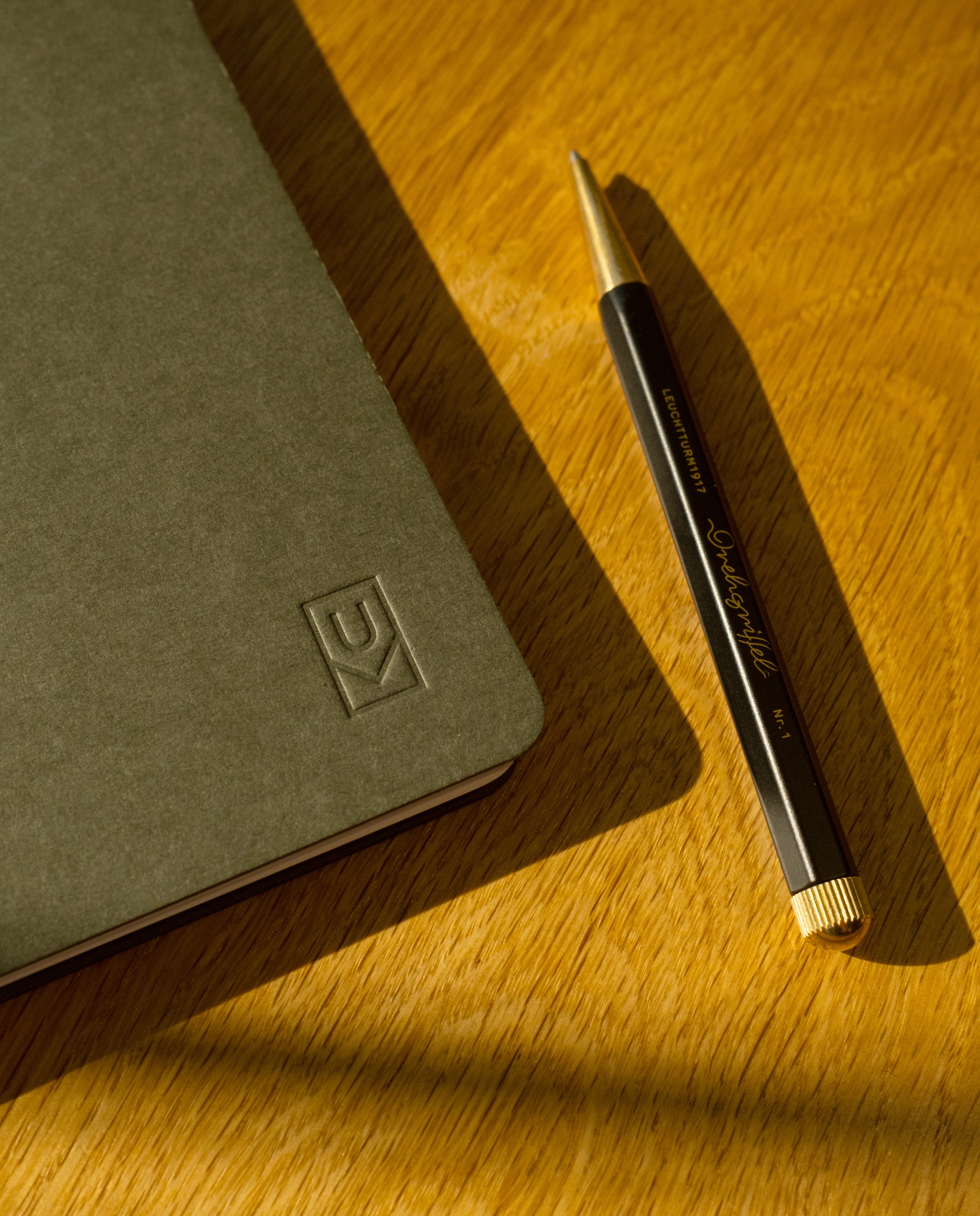 A black and gold pen rests on a wooden surface beside a closed, dark gray Discbound Journal (Standard 6"x 8.5") with an embossed logo in the corner, bathed in warm sunlight.