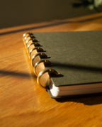 A close-up of the Discbound Journal (Standard 6"x 8.5") with solid brass discs, shown in dark colors on a wooden surface under warm lighting and soft shadows.