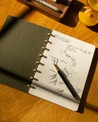 A Discbound Journal Bundle (Standard 6"x8.5" Journal + 3 Refills) with dot grid pages and sketches rests on a wooden desk, pen on the open page, sunlight playing across the paper, beside a wooden tray of pens and an hourglass.