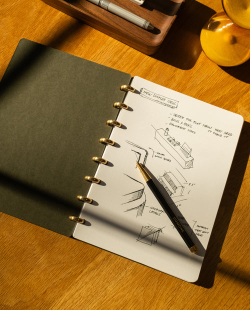 A Discbound Journal Bundle (Standard 6"x8.5" Journal + 3 Refills) with dot grid pages and sketches rests on a wooden desk, pen on the open page, sunlight playing across the paper, beside a wooden tray of pens and an hourglass.