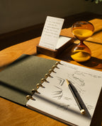 A Discbound Journal (Standard 6"x 8.5") with sketches and a pen rests on a wooden desk, next to an hourglass of yellow sand and an upright notepad with handwritten notes, all bathed in warm sunlight.