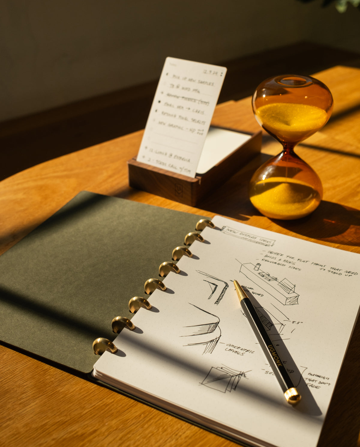A Discbound Journal (Standard 6"x 8.5") with sketches and a pen rests on a wooden desk, next to an hourglass of yellow sand and an upright notepad with handwritten notes, all bathed in warm sunlight.