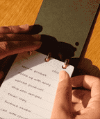 A hand holds a small notepad with a Discbound Analog Refill (6-pack) card on a wooden surface, featuring a to-do list with tasks such as reviewing a product, following up, and uploading products. Sunlight casts shadows across the page and hands, highlighting the convenience of using Discbound Analog Refill (6-pack) cards for daily organization.