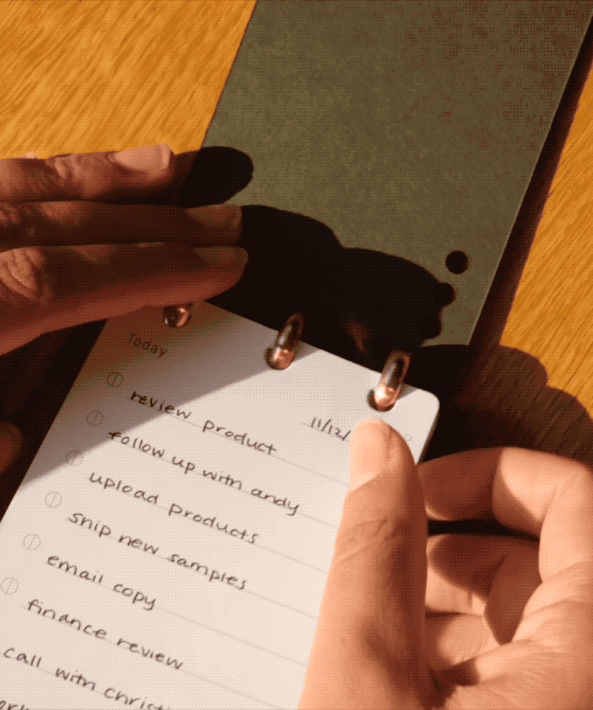 A hand holds a small notepad with a Discbound Analog Refill (6-pack) card on a wooden surface, featuring a to-do list with tasks such as reviewing a product, following up, and uploading products. Sunlight casts shadows across the page and hands, highlighting the convenience of using Discbound Analog Refill (6-pack) cards for daily organization.