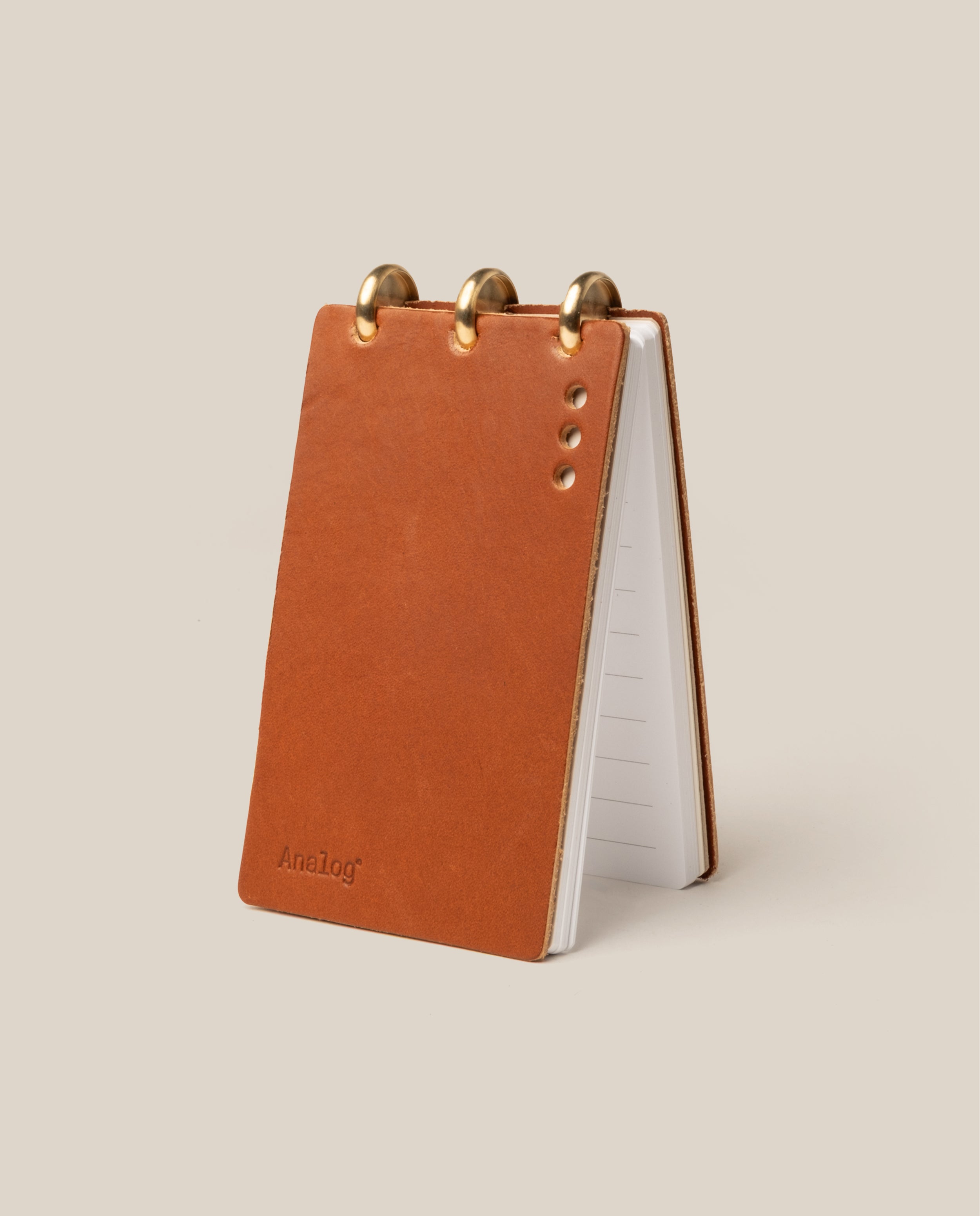 The Leather Discbound Analog (Tan) stands upright on a beige background, featuring a full-grain leather cover with four metal rivets, three brass rings, lined pages, and "Analog" embossed in the bottom corner.