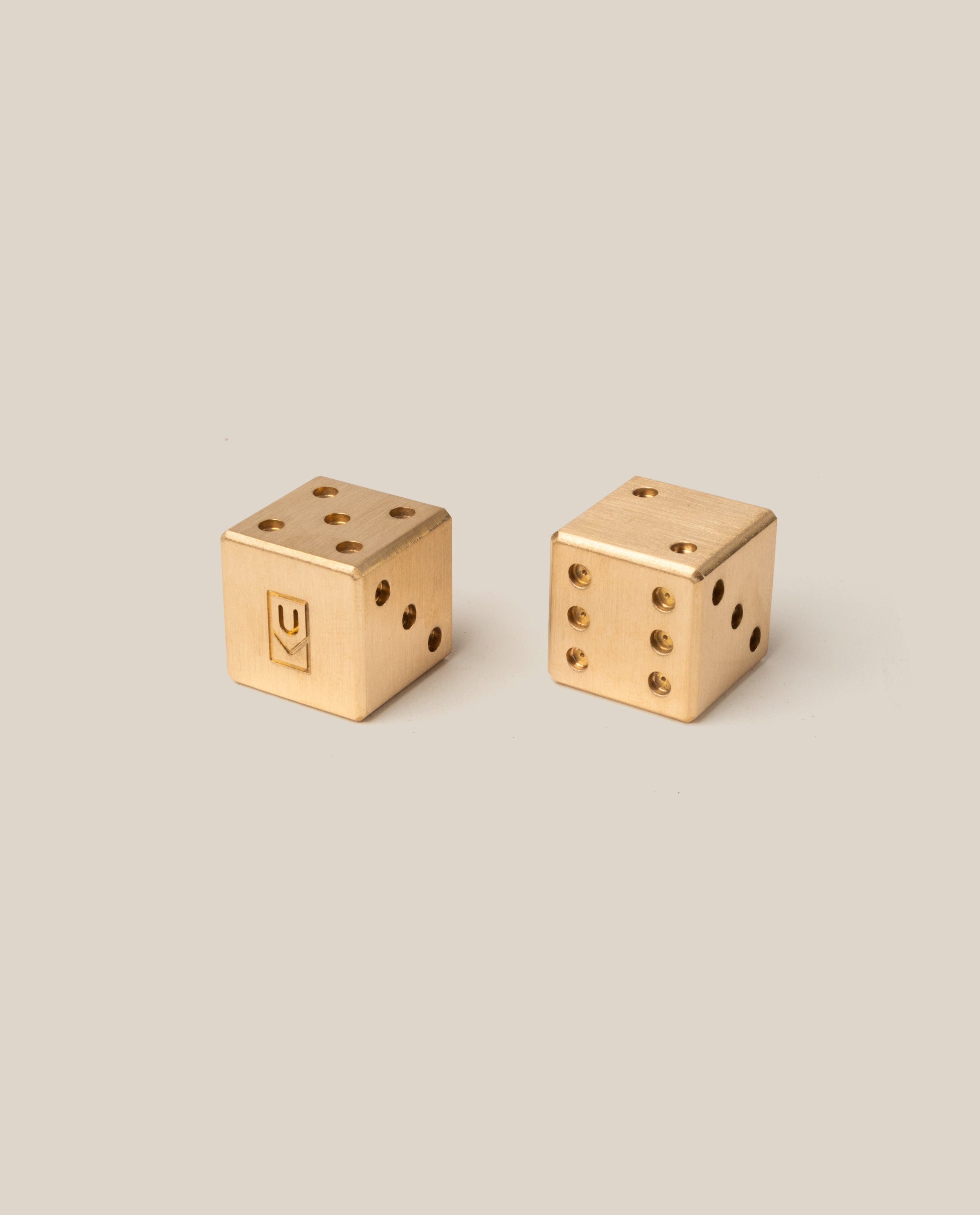 A pair of Ugmonk Brass Dice, crafted from gold-colored brass with precision milled detail, rest on a plain beige background; one die features Ugmonk’s signature symbol on a face in place of traditional pips, while the other displays classic dot patterns. These limited edition dice offer exquisite craftsmanship and a unique design, making them perfect for collectors and enthusiasts alike.