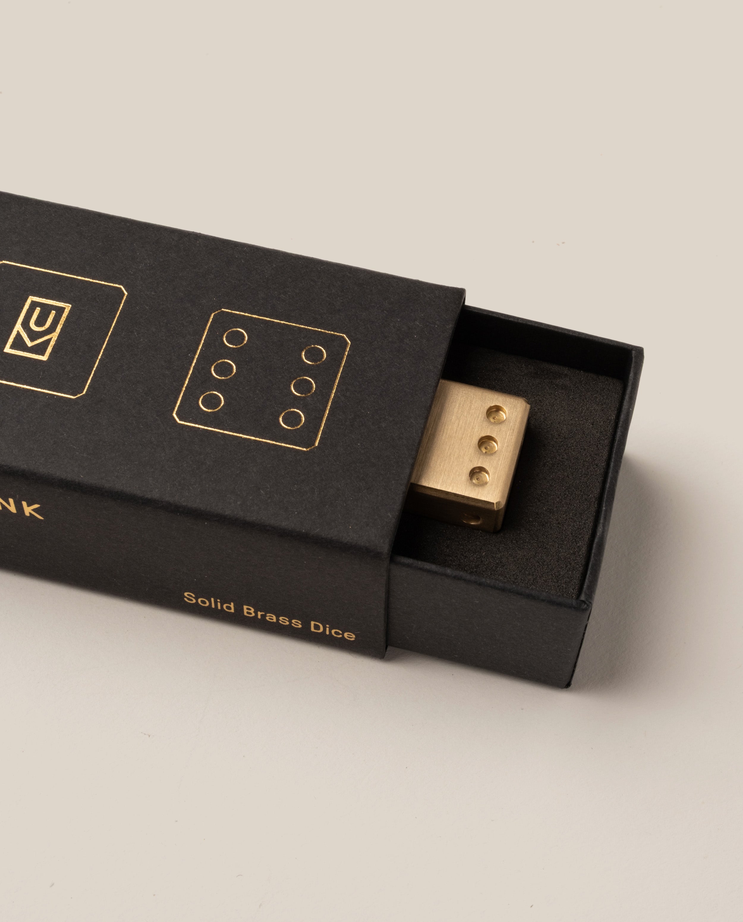 A precision-milled Ugmonk Brass Die partially slides out of a black box adorned with gold dice illustrations and text reading "Ugmonk Brass Dice" on the side. The visible face of the die displays four dots.