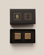Displayed on a neutral background, an open black box with matching gold designs on the lid reveals the Ugmonk Brass Dice: two gold-colored cufflinks—one adorned with an abstract symbol, and the other featuring six dots inspired by solid brass dice.