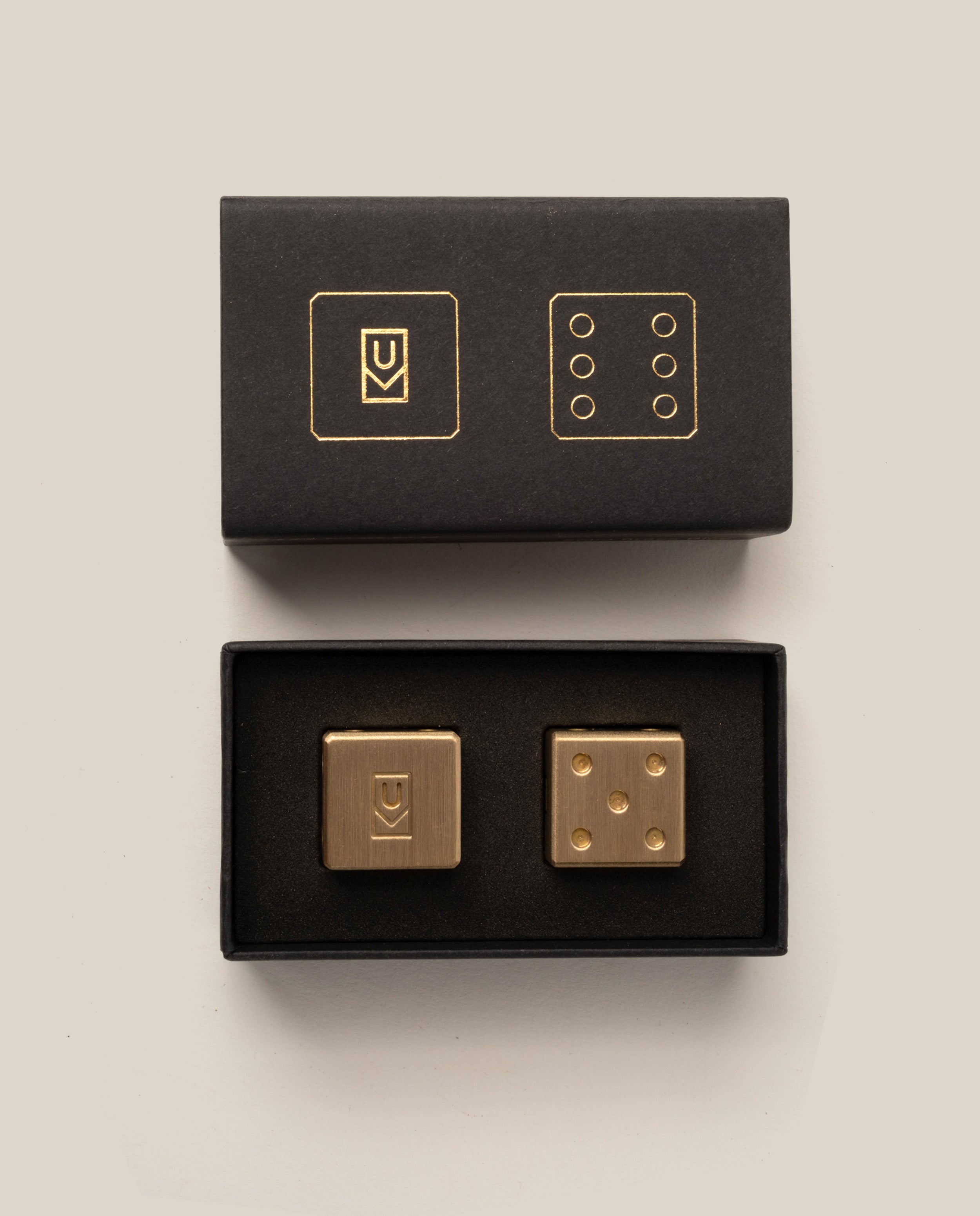 Displayed on a neutral background, an open black box with matching gold designs on the lid reveals the Ugmonk Brass Dice: two gold-colored cufflinks—one adorned with an abstract symbol, and the other featuring six dots inspired by solid brass dice.