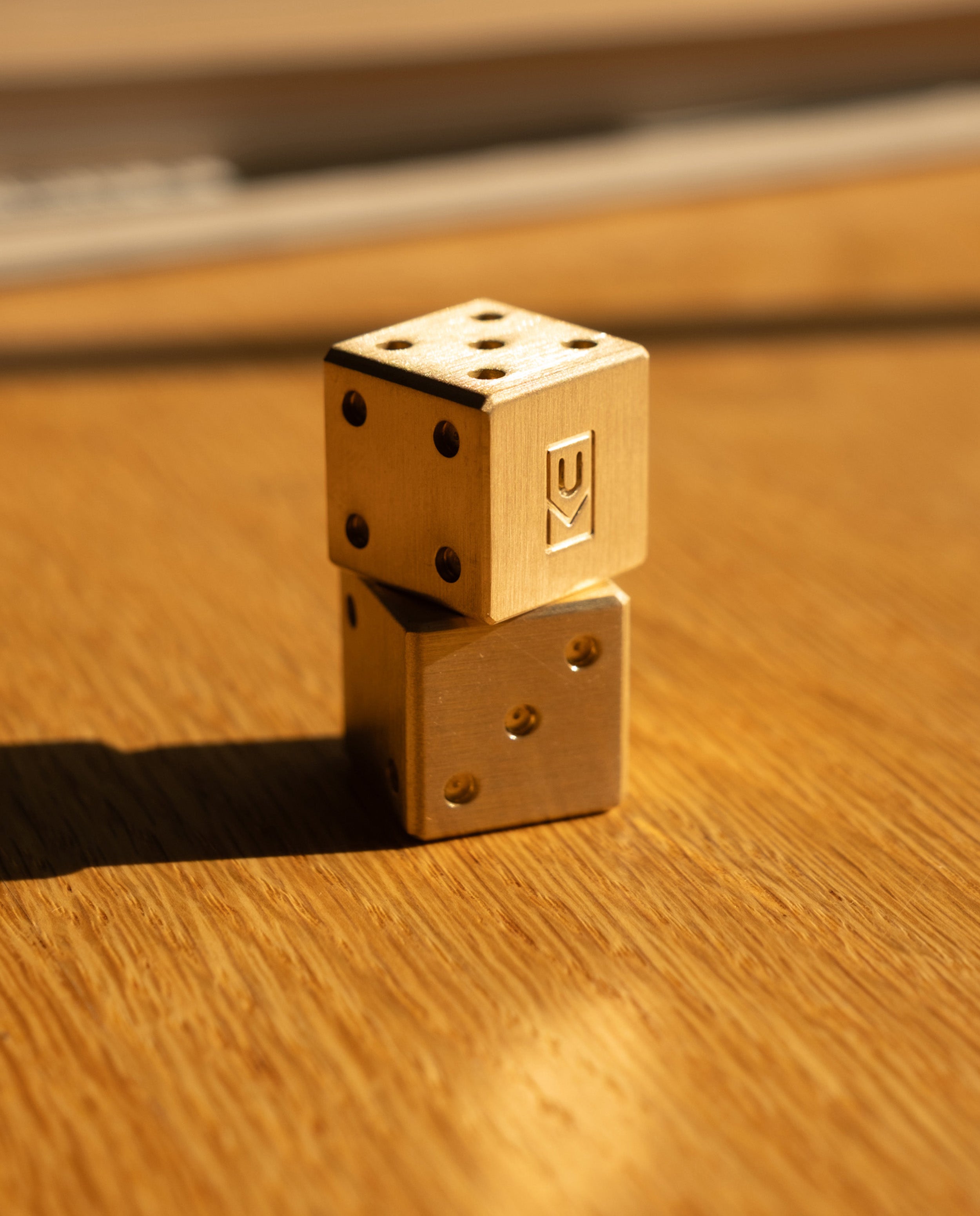 Two Ugmonk Brass Dice, precision-milled from solid brass, are stacked on a wooden surface bathed in warm sunlight. The top die reveals six dots and an engraved symbol, while the bottom die shows three dots. Shadows stretch to the left.