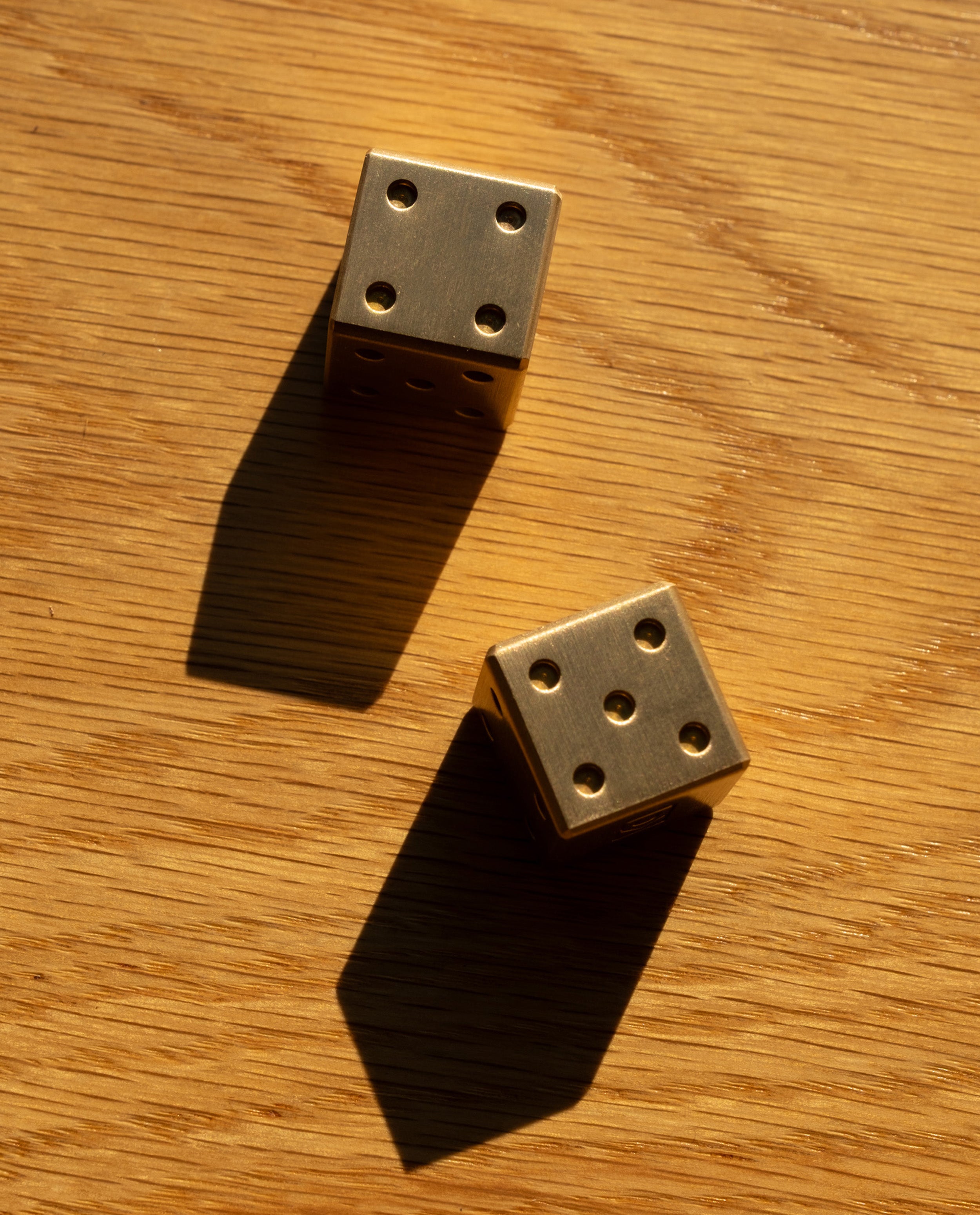 Two limited edition Ugmonk Brass Dice, displaying five and four, cast long shadows across a wooden surface in warm sunlight.