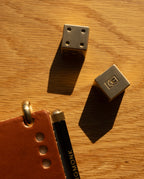 Two Ugmonk Brass Dice, precision-milled from solid brass, next to part of a brown leather notebook and a black pen on a wooden surface, illuminated by warm sunlight.
