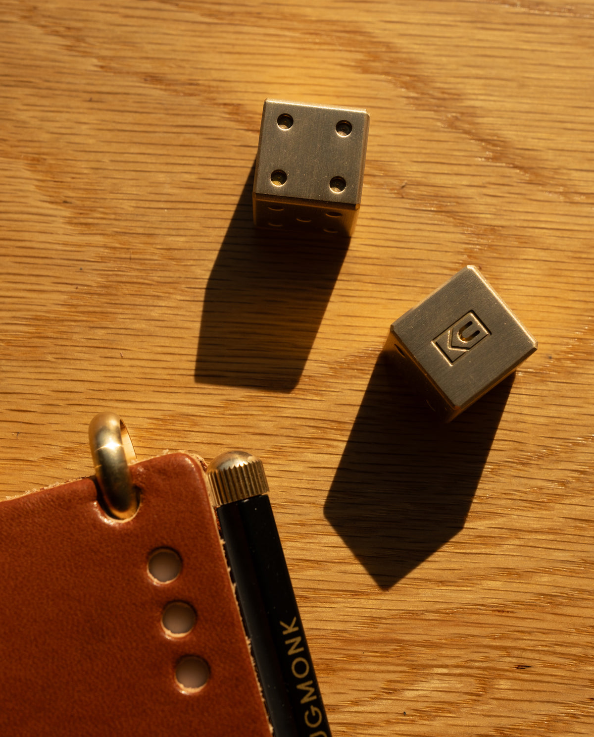 Two Ugmonk Brass Dice, precision-milled from solid brass, next to part of a brown leather notebook and a black pen on a wooden surface, illuminated by warm sunlight.