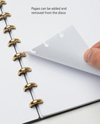 A hand lifts a Discbound Journal Refill - Dot Grid (Standard 6″ x 8.5″) page from a disc-bound planner with gold rings. Text reads, Pages can be added and removed from the discs, making it easy to customize your planner or Heirloom Discbound Journal with dot grid refill pages.
