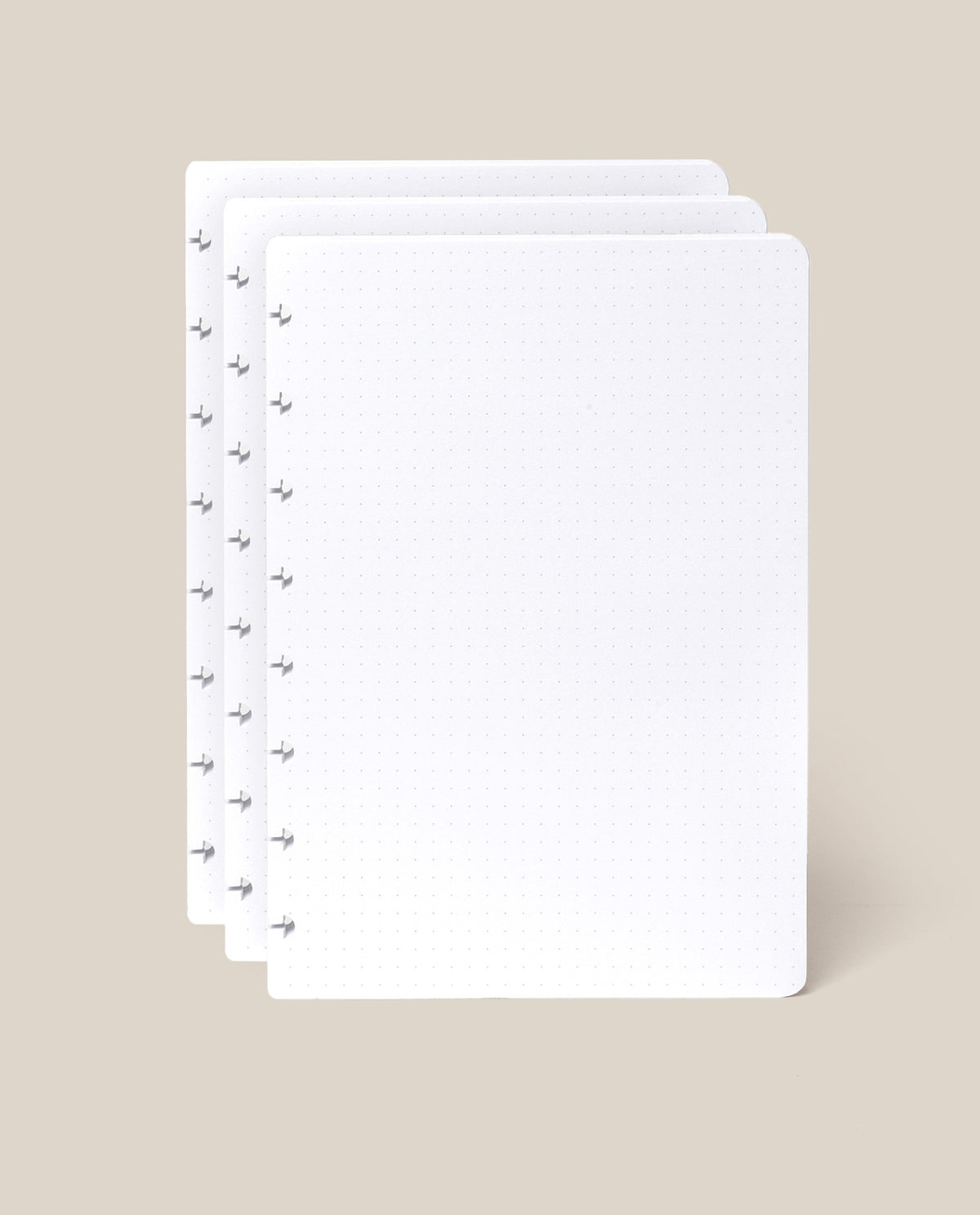 Three sheets of white, dot grid paper with rounded corners are stacked slightly offset on a beige background. The left side of each sheet features several small disc-binding holes, making them perfect as the Discbound Journal Refill - Dot Grid (Standard 6″ x 8.5″) for your Heirloom Discbound Journal or as part of a convenient journal refill pack.