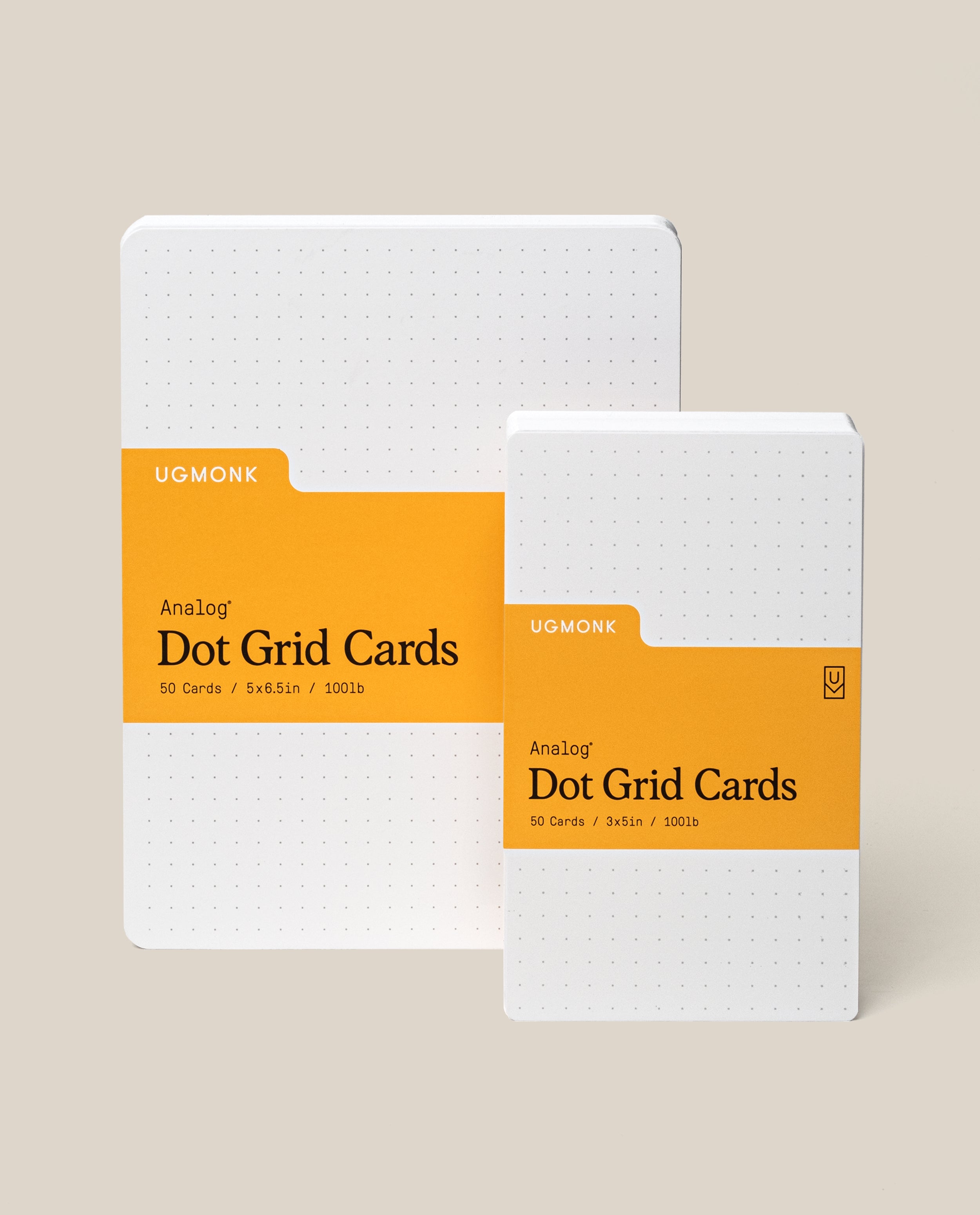 Two packs of Ugmonk Analog Dot Grid Cards, one large and one small, each with white dotted paper and an orange label displaying product details, standing upright on a beige background.