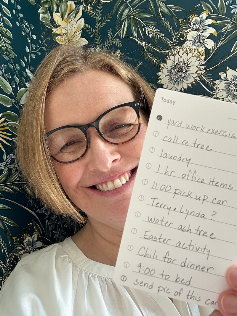A smiling person with glasses holds a handwritten to-do list in front of a floral wallpaper background.