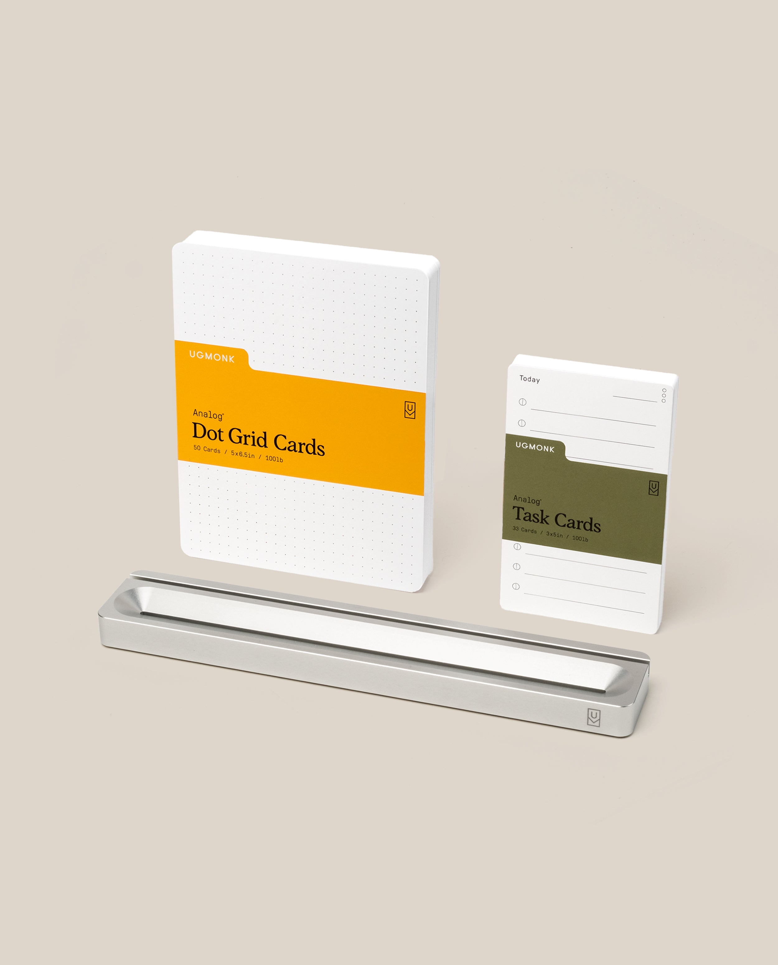 Two stacks of Ugmonk cards, one labeled Dot Grid Cards in yellow and the other Task Cards in green, are displayed upright behind a rectangular Analog Card Bar - Essentials Kit (Silver Aluminum) on a beige background.