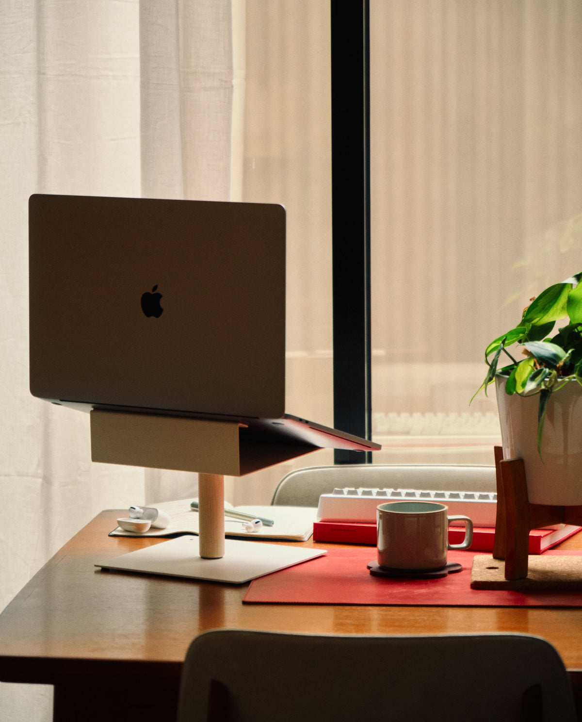 A Gather Laptop Stand (White/Maple) supports a laptop on a wooden desk by the window, alongside a keyboard, coffee cup, notebook, potted plant, and red placemat— all bathed in warm natural light.