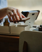 A person with dark nail polish lifts the Gather Solid Wood Lid (Walnut) from a small white organizer on a table, with a brown glass bottle and a digital device in the background.
