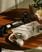 A wooden desk features headphones, a camera, pens, a smartphone on a stand, an open sketchbook, a small book, and magnetic Gather modules neatly arranged on the black Gather 1x4 Base Plate next to a checkered basket with a plant in sunlight.