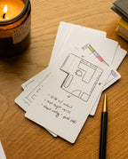 A stack of Analog - Graph Cards (3-Pack) with a hand-drawn floor plan and notes sits on a wooden desk beside a lit candle, a black and gold pen, and some stacked notebooks.