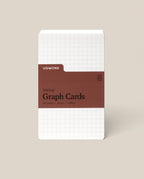 A pack of Analog Graph Cards - Small stands upright, made from uncoated cardstock with a white grid design and a brown label stating: Analog Graph Cards, 50 Cards, 3x5 in / 100 lb. The background is light beige.