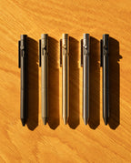 Five INVENTERY Bolt Action Pens (Zirconium)—a luxury writing instrument available in black, olive, gold, silver, and gunmetal—are neatly lined up on a wooden surface, casting soft shadows in bright natural light.