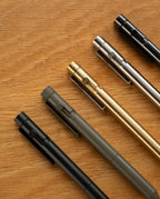 Five metal pens—including a sleek steel pen and the INVENTERY Bolt Action Pen (Onyx) finished with durable Cerakote ceramic coating—are arranged diagonally on a wooden surface in black, olive green, gold, silver, and matte black.