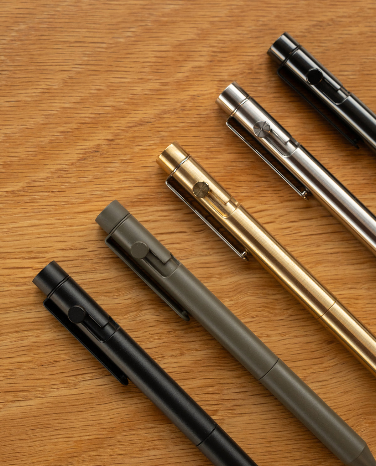 Five metal pens—including a sleek steel pen and the INVENTERY Bolt Action Pen (Onyx) finished with durable Cerakote ceramic coating—are arranged diagonally on a wooden surface in black, olive green, gold, silver, and matte black.