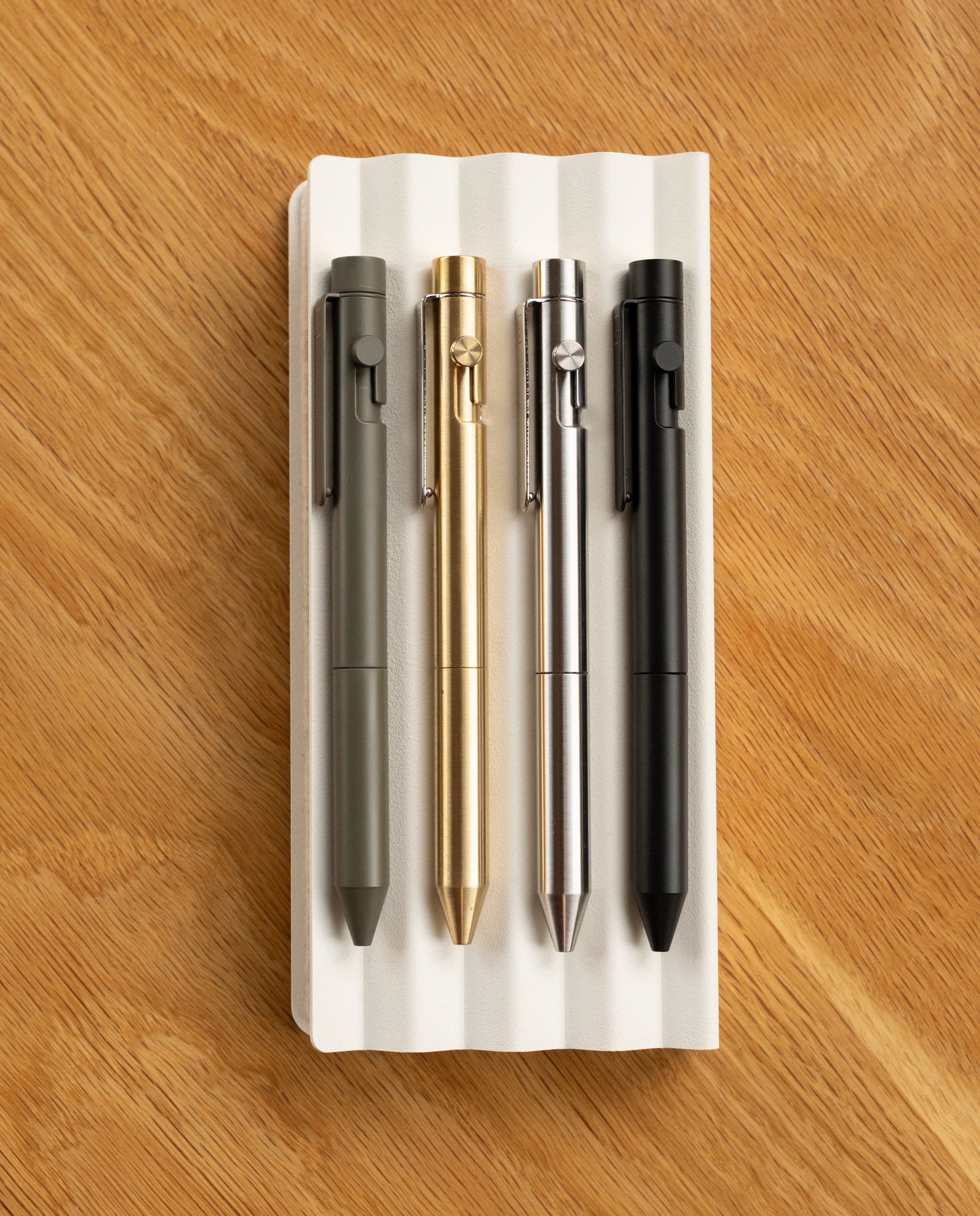 Four INVENTERY Bolt Action Pens (Stainless)—in green, gold, silver, and black—are lined up on a white grooved tray atop wood, each boasting a sleek, durable Cerakote finish.